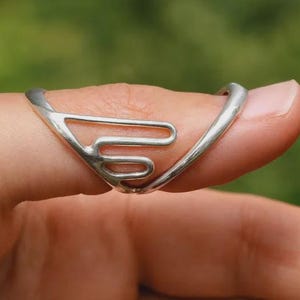 May include: A silver-colored ring with a unique, open design is worn on a finger. The ring's shape features a series of connected, curved lines, creating an abstract pattern. The ring is a piece of jewelry.