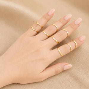 May include: A set of eight thin gold-toned rings worn on the fingers. The rings feature smooth, plain bands with a polished finish. Some fingers are adorned with two rings, while others feature a single ring, all showcasing a simple, uniform design.