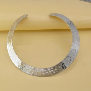Op de afbeelding: Een zilverkleurige metalen choker-halsketting met een breed, open ontwerp. Het oppervlak van de ketting heeft een opvallende gehamerde textuur, wat zorgt voor een reflecterend, ingedeukt uiterlijk over de gehele gebogen lengte van het sieraad.