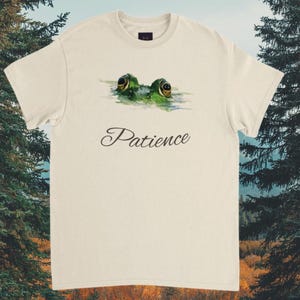 May include: Cream-colored t-shirt featuring a watercolor illustration of a frog's eyes peeking above the water. The word "Patience" is written in a cursive font below the frog image. The shirt has a crew neck and short sleeves.