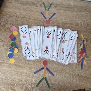 May include: A collection of educational toys, including colorful wooden sticks and circular tokens. The set features cards with stick figure designs and various colored shapes, ideal for learning and play. The toys are arranged on a light wood-grain surface.