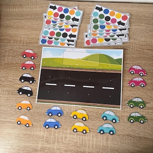 Pode incluir: Um jogo educativo infantil com uma cena rodoviária, uma estrada de asfalto preto, colinas verdes e um céu azul. Carros de brinquedo coloridos em várias cores estão espalhados, juntamente com cartões com setas e pontos coloridos.