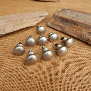 May include: Ten silver-toned cabinet knobs with a round, slightly domed shape. The knobs have a smooth, polished finish and are arranged in a scattered pattern on a brown burlap surface.