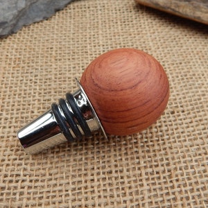May include: A wooden bottle stopper with a round, reddish-brown top and a silver metal base. The stopper has a black rubber ring around the base.