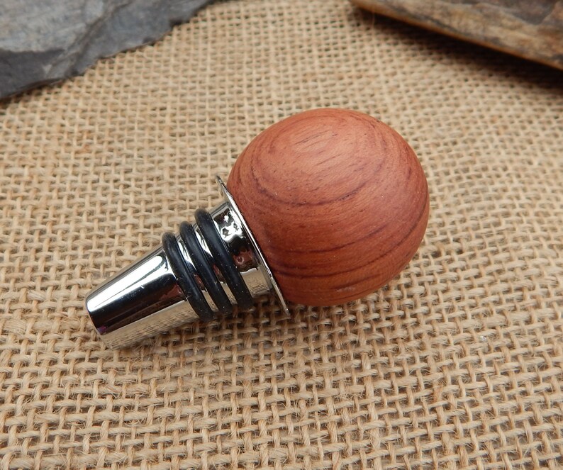 May include: A wooden bottle stopper with a round, reddish-brown top and a silver metal base. The stopper has a black rubber ring around the base.