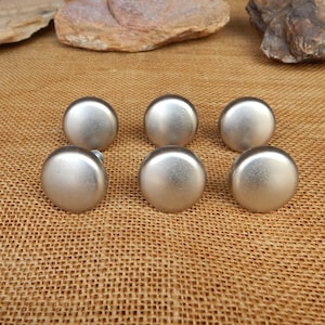May include: Six silver-colored cabinet knobs with a brushed metal finish. The knobs are round with a slightly domed top and a flat base. They are arranged in a group on a textured, tan surface.
