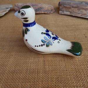 May include: A ceramic bird figurine with a speckled white body, adorned with blue and green floral designs. The bird has a blue collar, a green tail, and a green cap. The figurine is approximately 6 inches long.