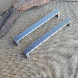 May include: Two rectangular cabinet pulls with a brushed silver finish. Each handle features a long, flat bar design with two perpendicular mounting posts at the ends, creating a clean, linear profile suitable for furniture or cabinetry hardware.