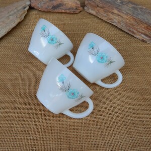 K&ouml;nnte beinhalten: Drei wei&szlig;e Keramikbecher mit blauen Blumendesigns. Die Becher haben eine abgerundete Form und einen Henkel an der Seite.