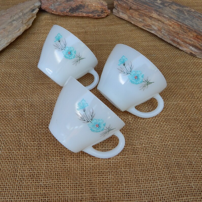K&ouml;nnte beinhalten: Drei wei&szlig;e Keramikbecher mit blauen Blumendesigns. Die Becher haben eine abgerundete Form und einen Henkel an der Seite.