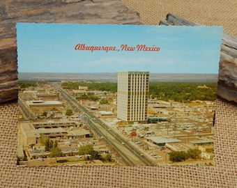 Peut inclure: Carte postale vintage représentant une vue aérienne d'Albuquerque, au Nouveau-Mexique. L'image montre un grand gratte-ciel blanc et beige, la First National Bank, dominant l'horizon de la ville. La carte postale a un bord festonné et les mots "Albuquerque, New Mexico" en rouge.