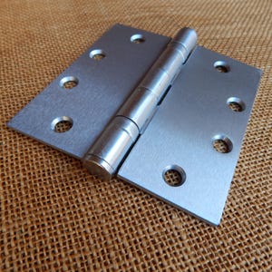 May include: A silver door hinge with five barrel-shaped pins. The hinge is made of metal and has a brushed finish.