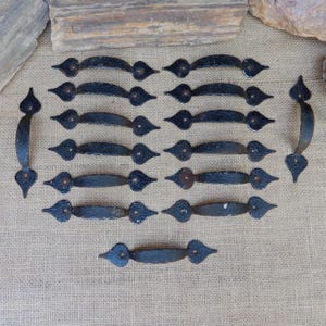 May include: A collection of thirteen black metal drawer pulls with a textured finish. Each pull features a curved handle and pointed, spade-shaped ends with screw holes. The set includes eleven horizontal pulls and two vertical pulls arranged in a grid.
