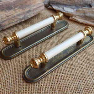 May include: Two antique door handles. Each handle has a rectangular, bronze-colored base with a white ceramic bar and gold-colored accents. The handles are designed for a classic or vintage aesthetic.