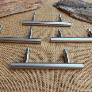 May include: Five brushed nickel cabinet pulls with a rectangular shape and rounded edges. The pulls have a simple, modern design.
