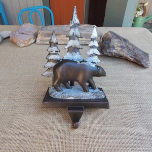 May include: A bronze-colored bear figurine walks past snow-covered pine trees. The decorative piece is designed to hold a Christmas stocking. The base is rectangular and has a small hook at the bottom.