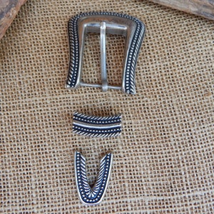 K&ouml;nnte beinhalten: Silberne G&uuml;rtelschnalle mit dekorativem Design. Die Schnalle ist aus Metall und hat eine schwarze Oberfl&auml;che. Die Schnalle besteht aus drei Teilen, einer Hauptschnalle, einem Riemenhalter und einer Spitze.