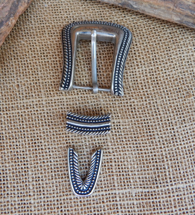 K&ouml;nnte beinhalten: Silberne G&uuml;rtelschnalle mit dekorativem Design. Die Schnalle ist aus Metall und hat eine schwarze Oberfl&auml;che. Die Schnalle besteht aus drei Teilen, einer Hauptschnalle, einem Riemenhalter und einer Spitze.