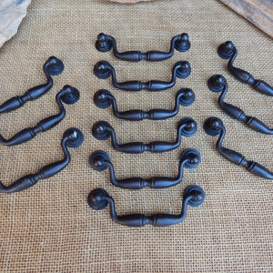 May include: A set of 12 black metal cabinet pulls with a curved design. The pulls are arranged in rows on a burlap background.