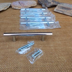 May include: A collection of polished chrome cabinet pulls with matching screws. The pulls have a cylindrical bar design with two round posts for mounting. Several pulls are packaged in clear plastic, with a single pull and screws displayed separately.
