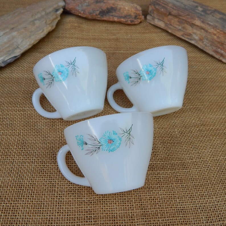K&ouml;nnte beinhalten: Drei wei&szlig;e Keramikbecher mit blauen Blumendesigns. Die Becher haben eine abgerundete Form und einen Henkel an der Seite.