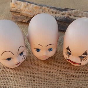 May include: Three porcelain doll heads with painted faces. The doll heads have blue eyes, painted eyebrows, and lips. One doll head has a painted tear on its cheek.