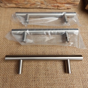 May include: Stainless steel cabinet pulls. The pulls are cylindrical with a brushed finish. Two pulls are in clear plastic packaging. The pulls are on a burlap surface.