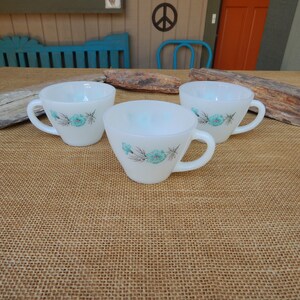 K&ouml;nnte beinhalten: Drei wei&szlig;e Keramikbecher mit blauen Blumendesigns. Die Becher haben eine abgerundete Form und einen Henkel an der Seite. Die Becher stehen auf einer braunen Jutestruktur.