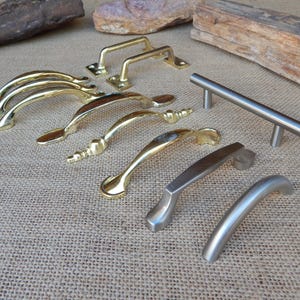 May include: A collection of cabinet and drawer pulls in various styles and finishes. The hardware includes gold, brushed nickel, and stainless steel options. The pulls are arranged on a textured, neutral-toned surface.