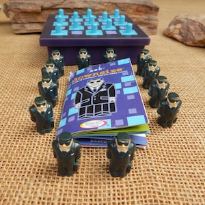 May include: A board game called "downsize" with a purple game board and light blue pegs. The game includes small figurines of men in black suits and sunglasses. The game's box is purple and features a cartoon of a man in a suit.