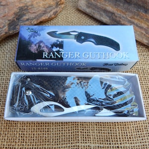 May include: A black folding knife with a gut hook blade. The knife is in a white box with the words "Ranger Guthook" printed on it. The box is on a brown burlap surface.