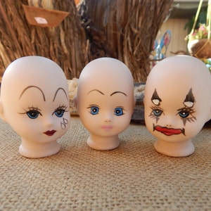 May include: Three porcelain doll heads with painted faces. The doll heads have blue eyes, painted eyebrows, and red lips. One doll head has a clown-like makeup design.