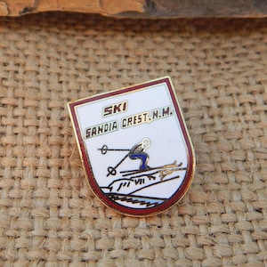 May include: A gold and red enamel pin with a white background. The pin features a skier in blue and white clothing, skiing down a mountain. The text "SKI SANDIA CREST N.M." is printed in gold on the pin.