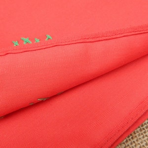 May include: Close-up of folded coral-colored fabric with a visible seam and green embroidered details. The fabric is smooth and appears to be a lightweight material, possibly cotton or a blend. The stitching is visible along the edge.