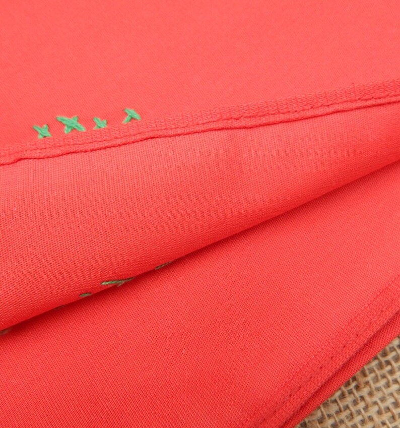 May include: Close-up of folded coral-colored fabric with a visible seam and green embroidered details. The fabric is smooth and appears to be a lightweight material, possibly cotton or a blend. The stitching is visible along the edge.