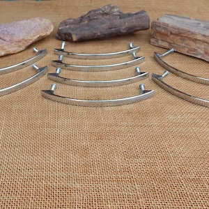 May include: A set of ten silver-toned, arched cabinet pulls. The pulls are arranged in two rows on a brown burlap surface.