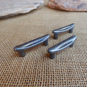 May include: Three silver metal drawer pulls with a curved, slightly rounded shape. The pulls are arranged in a row on a brown burlap surface.