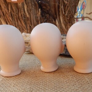 May include: Three unpainted porcelain doll heads, each with a smooth, light pink finish. The heads are positioned side-by-side, facing the right, with their backs visible.
