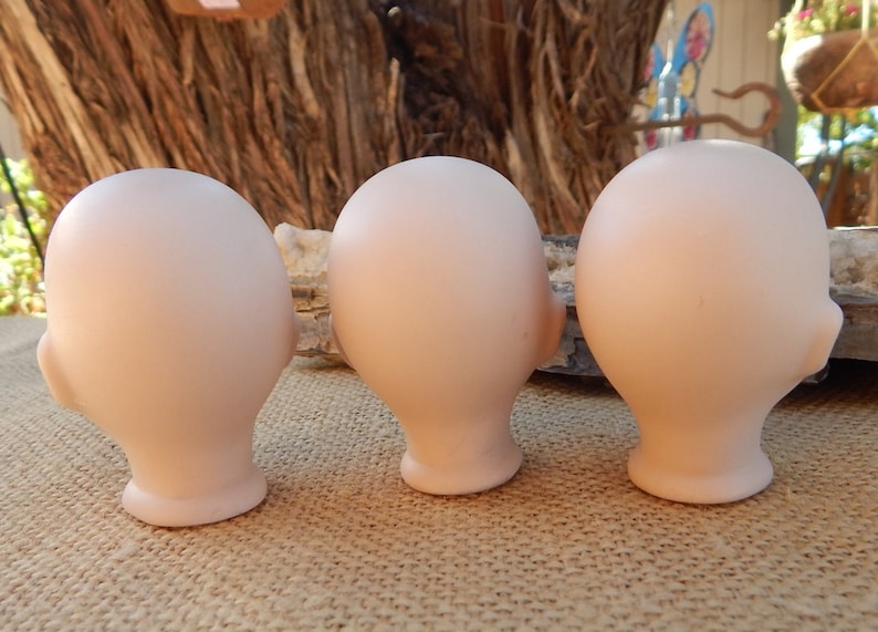 May include: Three unpainted porcelain doll heads, each with a smooth, light pink finish. The heads are positioned side-by-side, facing the right, with their backs visible.