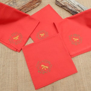 May include: Four red cloth napkins with embroidered gold wreaths and bows. The napkins are square and folded, with the embroidered design in the center of each. The napkins are arranged on a textured, neutral-colored surface.