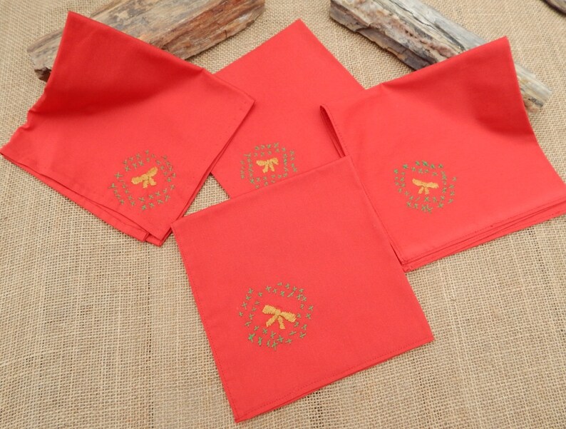May include: Four red cloth napkins with embroidered gold wreaths and bows. The napkins are square and folded, with the embroidered design in the center of each. The napkins are arranged on a textured, neutral-colored surface.