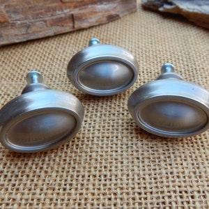 May include: Three silver oval-shaped cabinet knobs with a brushed finish. The knobs have a raised center and a smooth, rounded edge.
