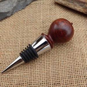 K&ouml;nnte beinhalten: Ein Flaschenverschluss aus Holz mit dunkelroter Oberfl&auml;che und einem verchromten Metallfu&szlig;. Der Verschluss hat eine Gummidichtung, um Lecks zu verhindern.