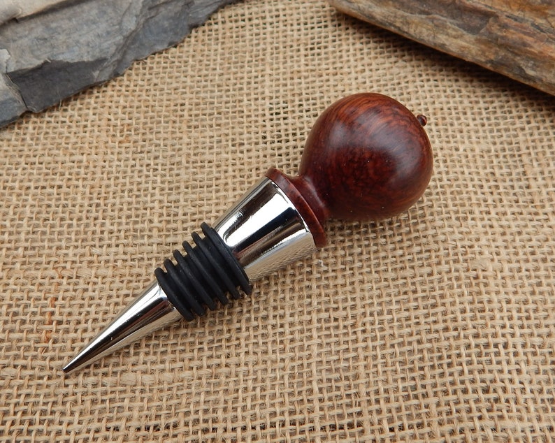 K&ouml;nnte beinhalten: Ein Flaschenverschluss aus Holz mit dunkelroter Oberfl&auml;che und einem verchromten Metallfu&szlig;. Der Verschluss hat eine Gummidichtung, um Lecks zu verhindern.