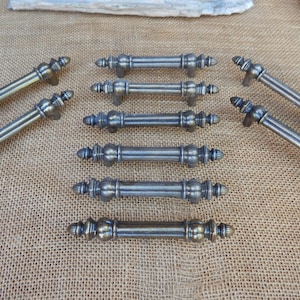 May include: A collection of antique-style bronze cabinet pulls. The pulls have a detailed design with decorative ends and a central, rounded section. The hardware is arranged on a textured, tan surface, suggesting a rustic or vintage aesthetic. Ideal for furniture restoration.