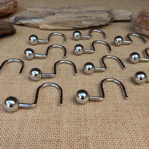 May include: A collection of shiny, chrome-colored shower curtain hooks. Each hook has a spherical ball at one end and a curved, open hook shape. The hooks are arranged on a textured, tan surface, with pieces of wood in the background.