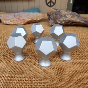 May include: Six silver-colored cabinet knobs with a geometric, multi-faceted design. Each knob has a rounded base and a pentagonal top. The knobs are arranged on a textured, tan surface.