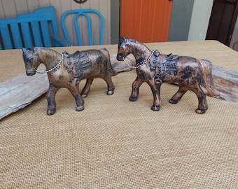 May include: Two small, black and copper colored metal horse figurines with saddles. The horses are standing on a brown burlap surface.