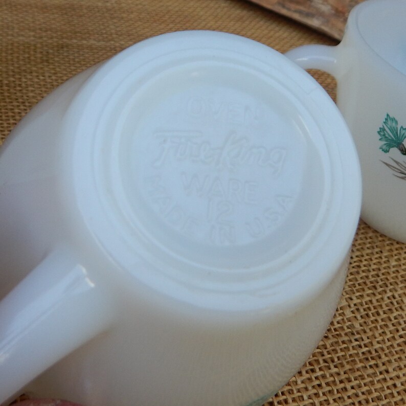 K&ouml;nnte beinhalten: Wei&szlig;e Keramiktasse mit gr&uuml;nem Blumendesign und einem "Fire King Ware" Logo auf der Unterseite.
