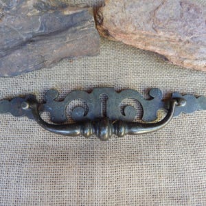May include: A dark metal drawer pull featuring a decorative backplate with an intricate scrollwork design and a central keyhole cutout. The handle is a curved, swinging bail style with a rounded center grip, finished in a muted, aged metallic tone.